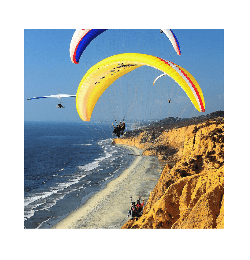 Multiple paragliders flying over a beach with cliffs, ocean waves, and clear blue sky.