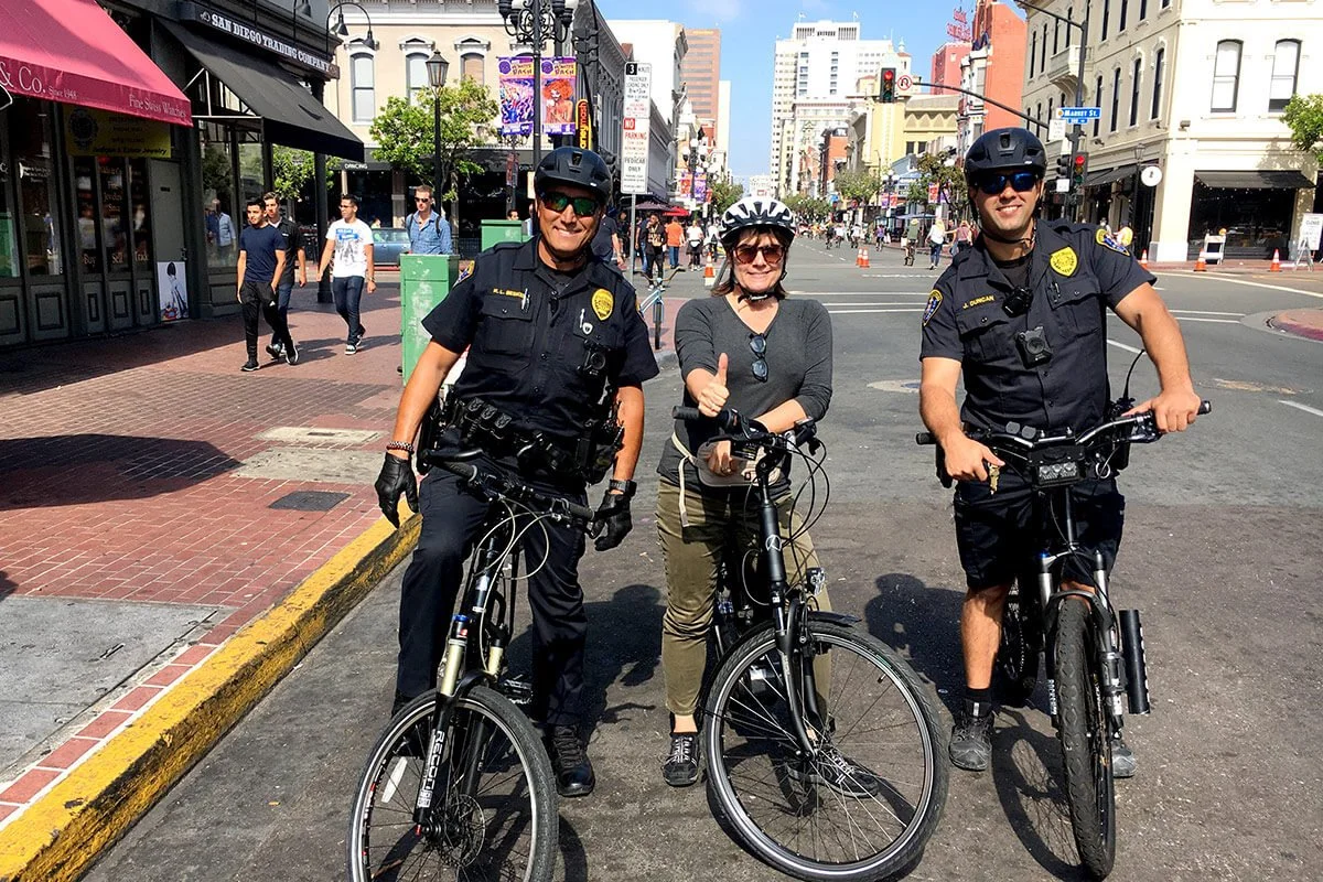 Two police officers and a woman posing with bicycles on a city street, with pedestrians and buildings in the background.