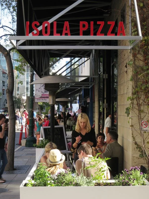 Outdoor scene at Isola Pizza with customers dining and walking on the sidewalk.