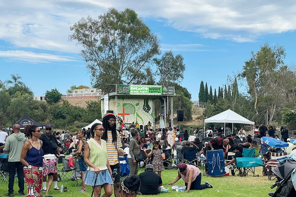 Crowd enjoying a music festival outdoors, with people sitting on the grass, standing, and dancing in front of a stage with a large screen, trees, and tents in the background.
