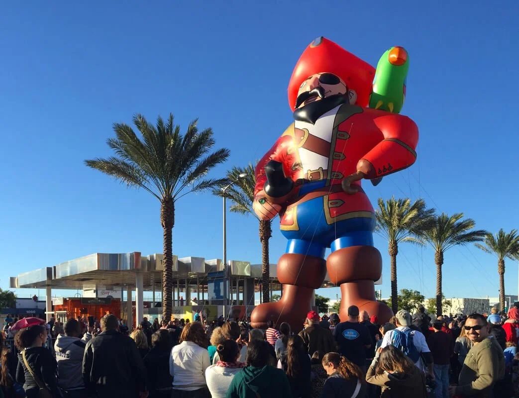 Large inflatable pirate with a red bandana, black eye patch, red coat, and blue pants, standing among palm trees and a crowd of people.