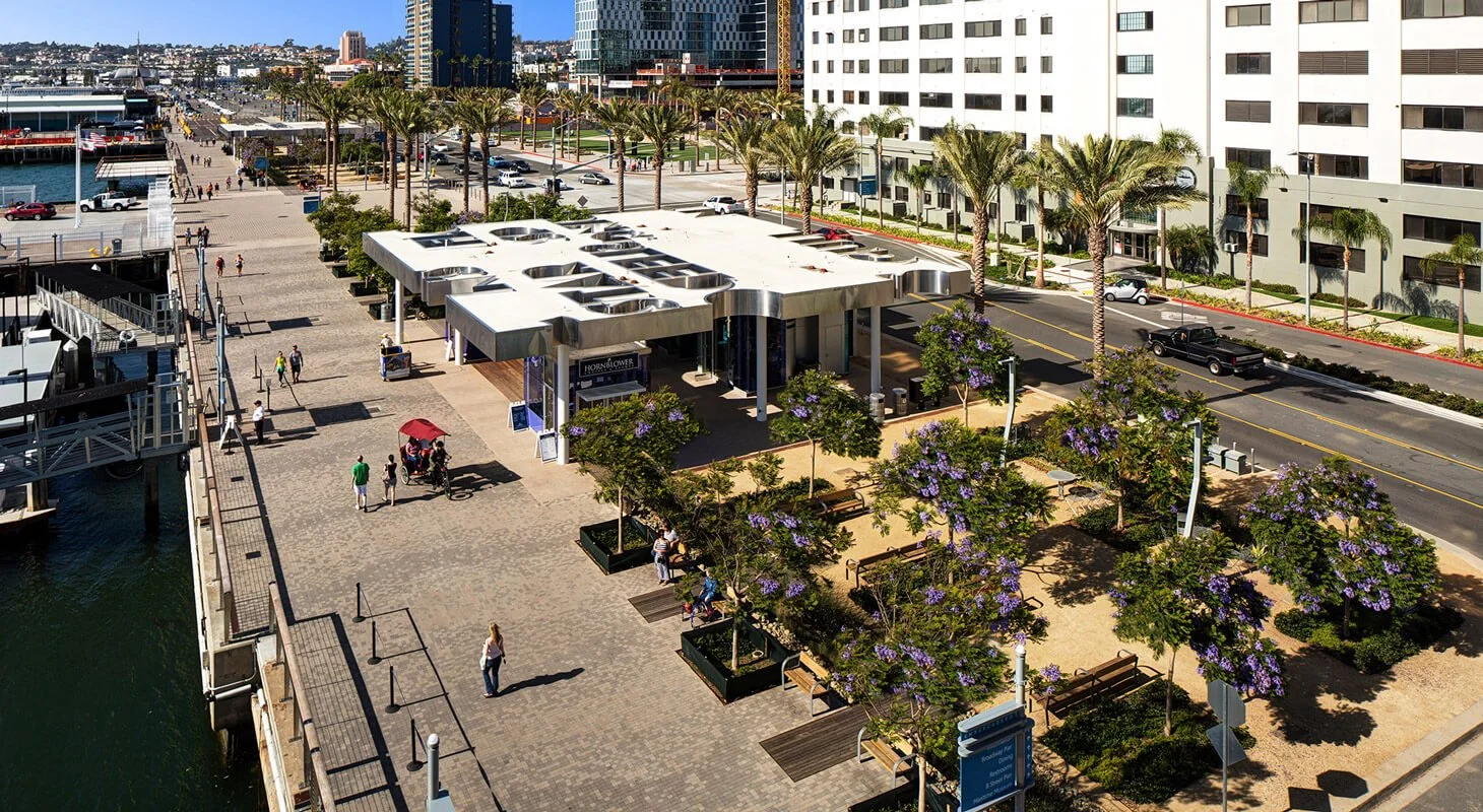 Aerial view of a waterfront promenade with trees, benches, and a modern white building, with a street and high-rise buildings in the background.