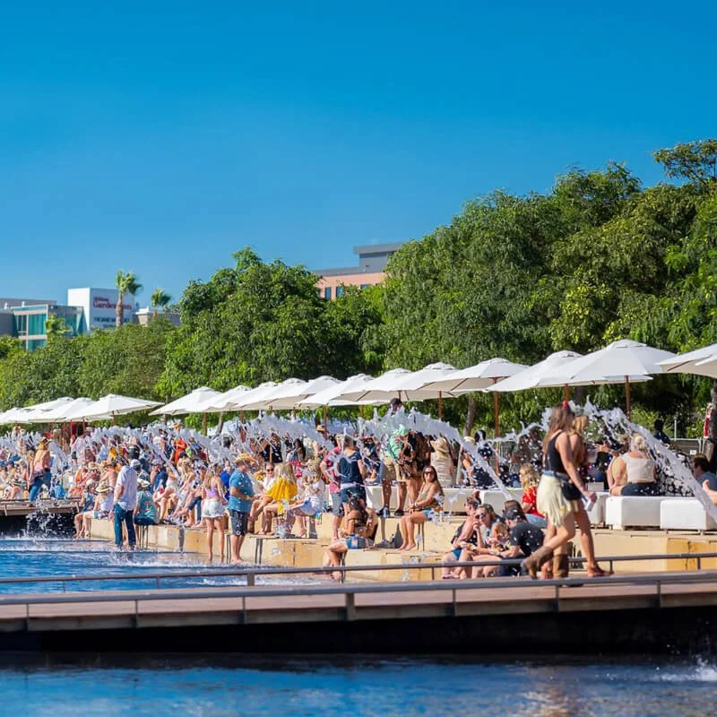 Crowded beach scene with many people sitting under white umbrellas, water splashing, green trees, and modern buildings in the background on a sunny day.
