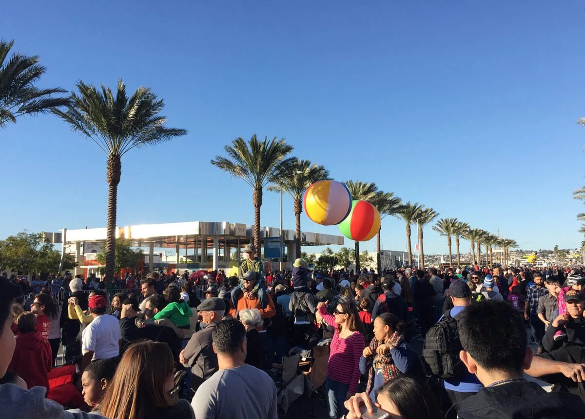 Large crowd of people gathered outdoors with palm trees and a modern building in the background, under a clear blue sky.
