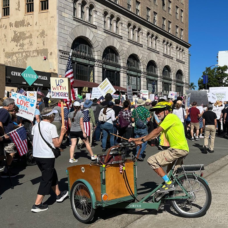 A crowd of people participating in a protest or rally on a city street with many holding American flags and signs. A man in a yellow shirt and helmet is riding a green cargo bicycle in the foreground.