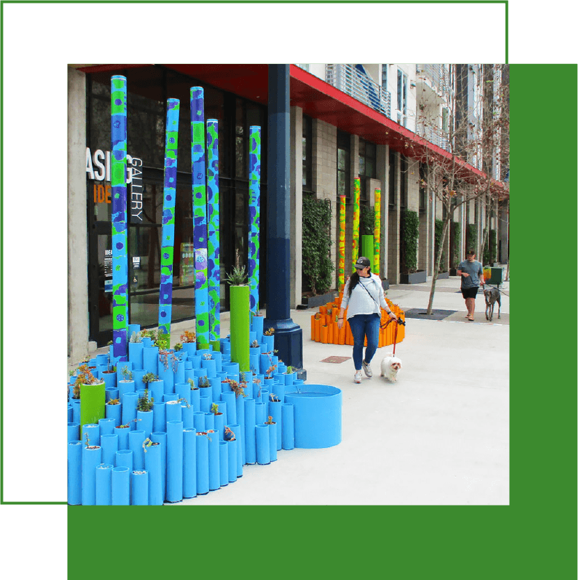 Outdoor mural sculpture with blue PVC pipes and colorful vertical poles next to a gallery entrance on a city sidewalk, with two people walking dogs in the background.