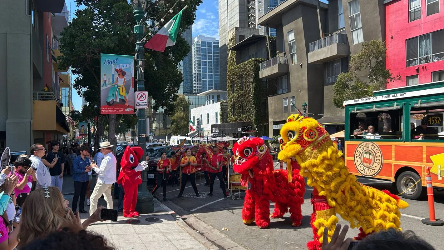 Two lions in colorful costumes performing on a city street during a parade, with onlookers taking photos and a trolley in the background.