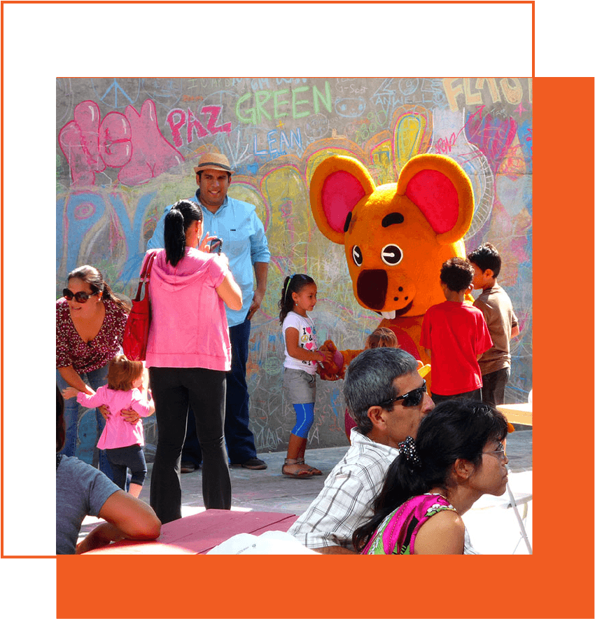 Children and adults at a park with a person in a large mouse costume, with colorful graffiti on a wall behind them.