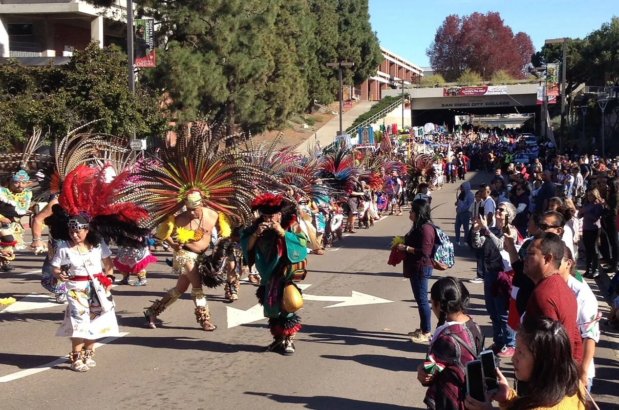 A parade featuring dancers in colorful costumes with feathers, and a crowd of spectators along a city street.