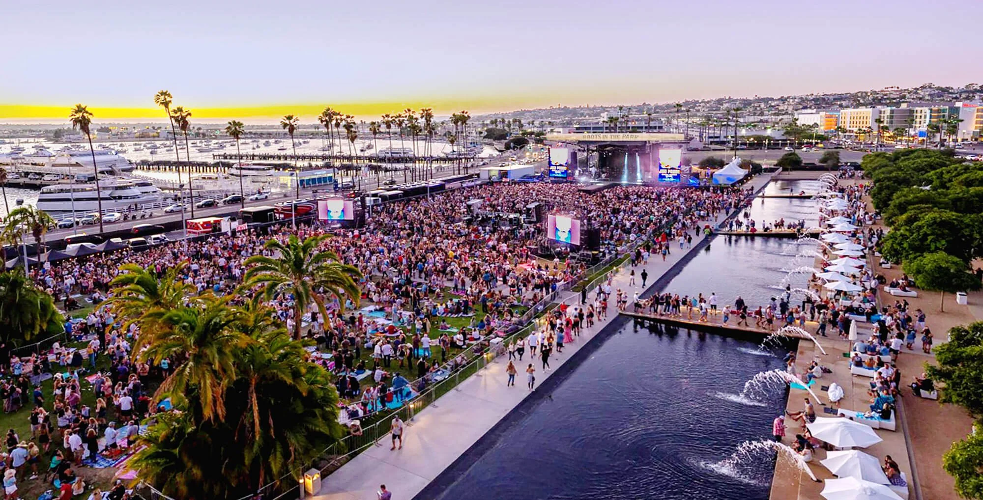 An outdoor concert at a marina with a large crowd gathered on the grass and around water features, palm trees, yachts, and boat docks in the background at sunset.