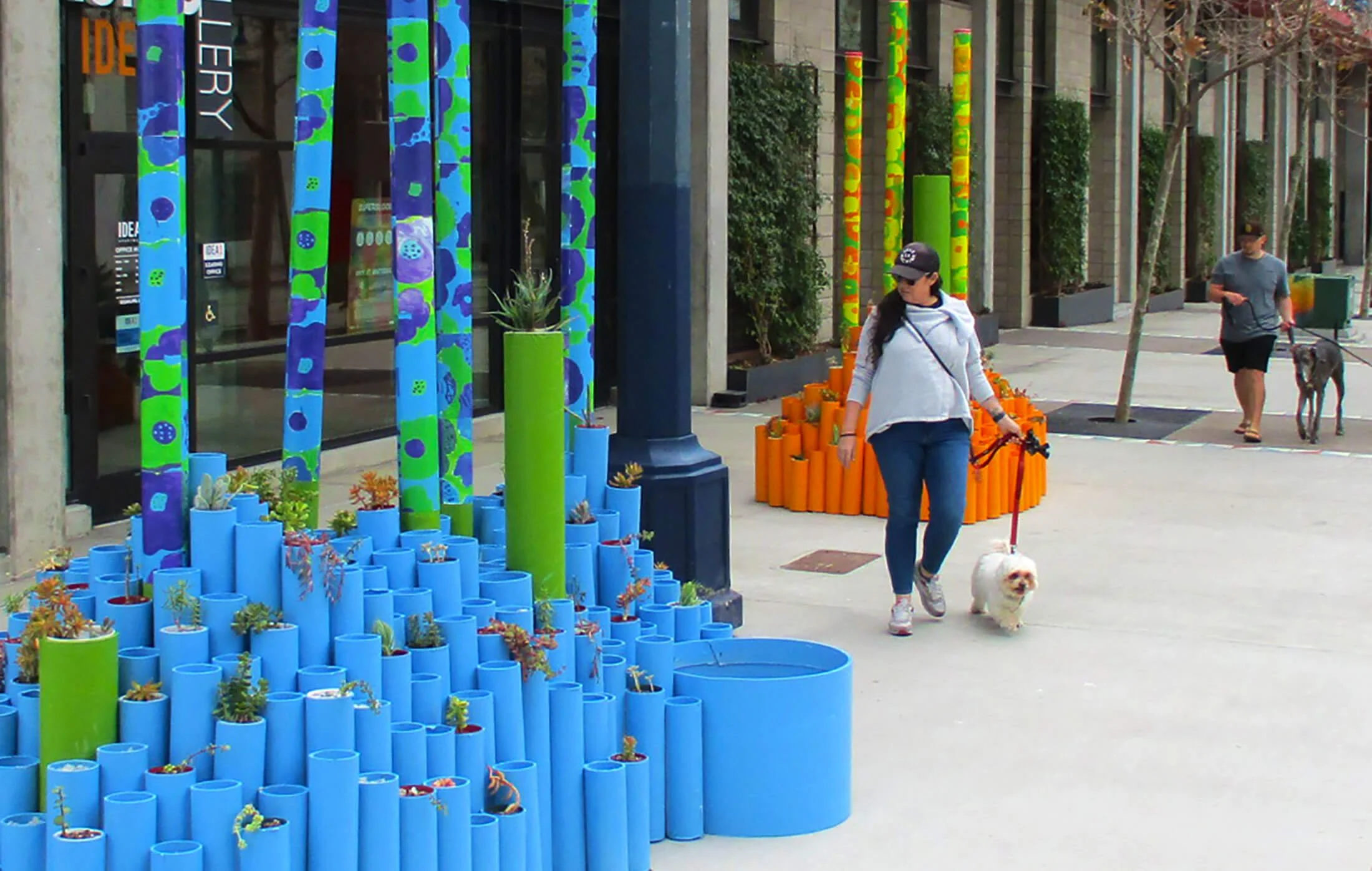 Colorful outdoor art installation with vertical and circular painted pipes, plants, and people walking with dogs on a city sidewalk.