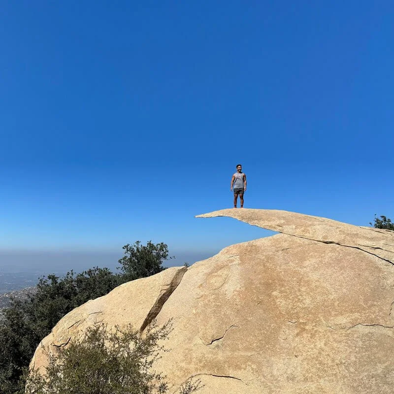A person standing on a small flat rock ledge extending from a large boulder, with a clear blue sky in the background and some trees below.