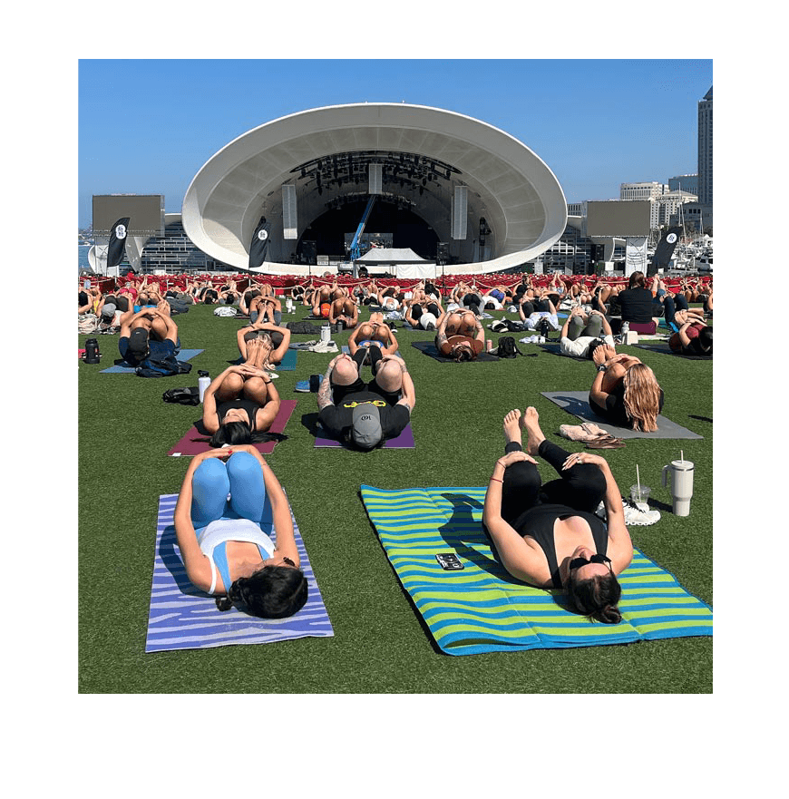 People practicing yoga outdoors on colorful mats on a grassy area near a stage with a curved white roof, against a city skyline under a clear blue sky.