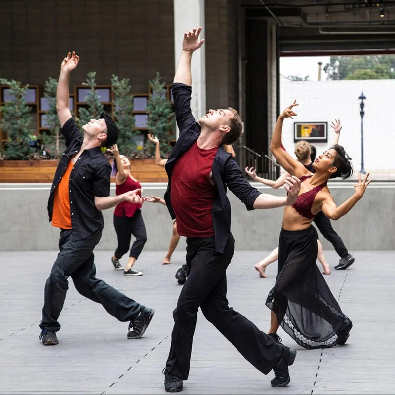 A group of dancers performing in an outdoor space, with some dancers reaching upward and others in expressive poses.