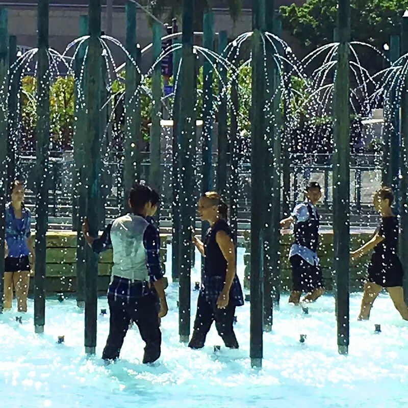 Children playing in a splash pad with water sprays and foam, surrounded by trees and a green fence.