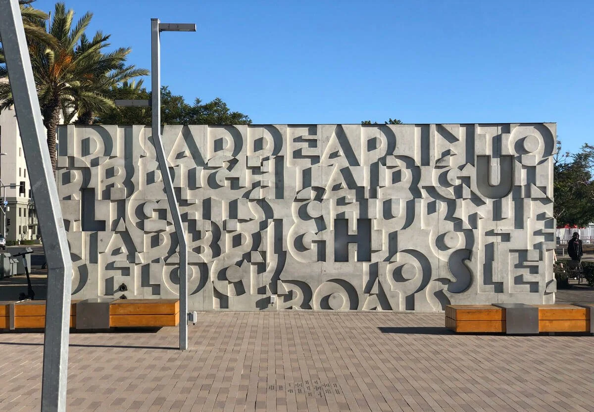 Large concrete wall with cut-out letters spelling 'DISAPPEAR TO REBEL AGAIN LAUGH AND GUILT HAVE BEEN LEFT BEHIND' in a public space with benches, trees, and a blue sky.