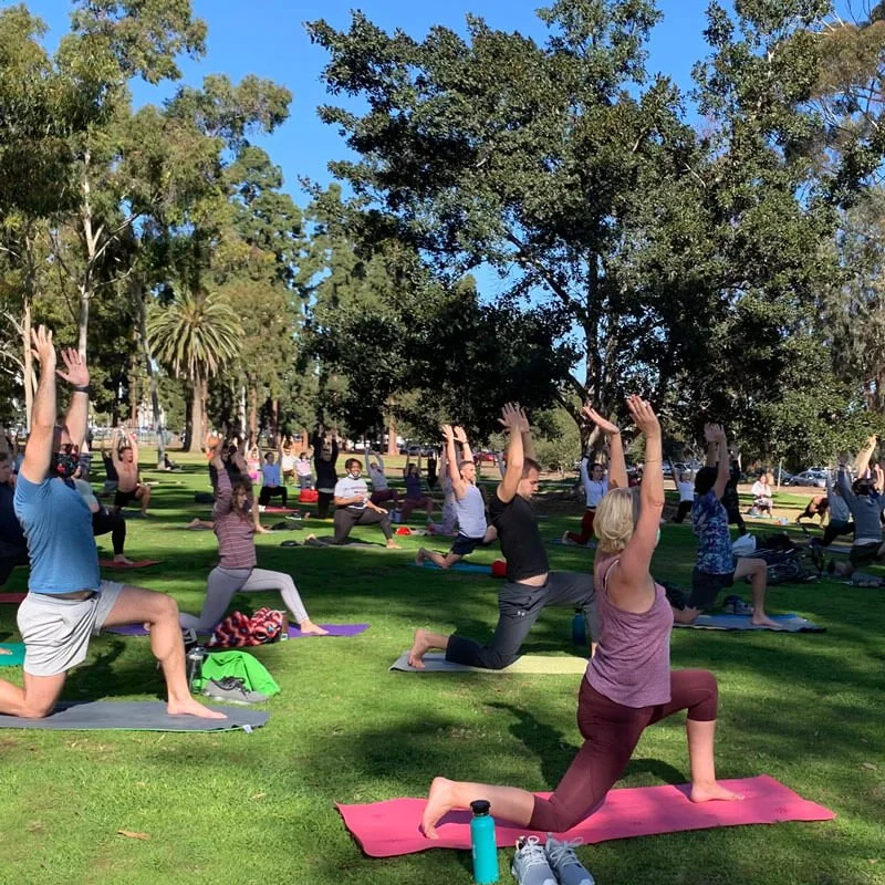 Group of people practicing yoga outdoors on a sunny day in a park, standing on yoga mats with trees in the background.