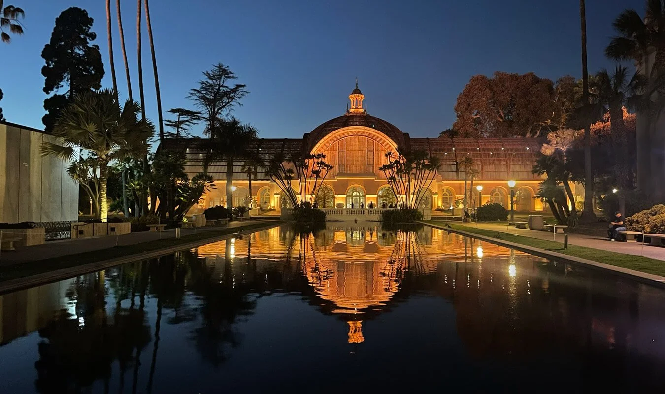 Night view of a historic greenhouse building with arch windows, illuminated and reflected in a long rectangular water feature, surrounded by trees and park benches.