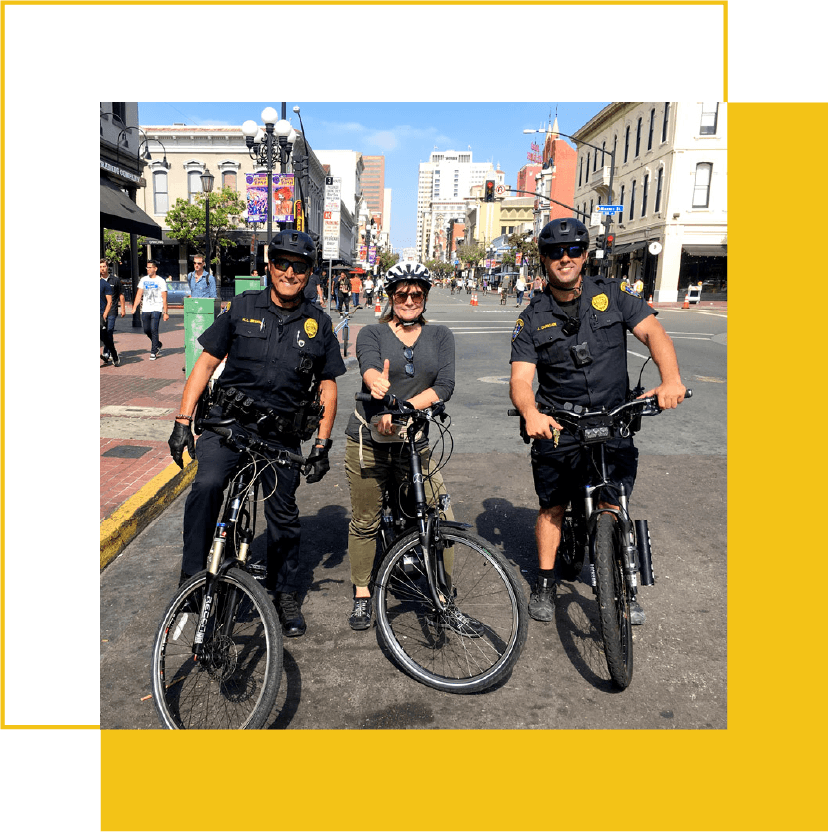 A woman standing between two police officers on bicycles in a city street, giving a thumbs-up while wearing a helmet, sunglasses, and casual clothes on a sunny day.