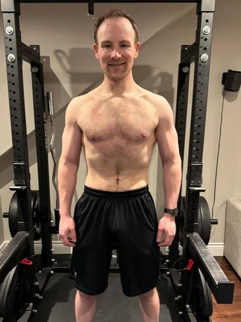 A man standing shirtless in a gym, smiling, with a power rack and weight plates in the background.
