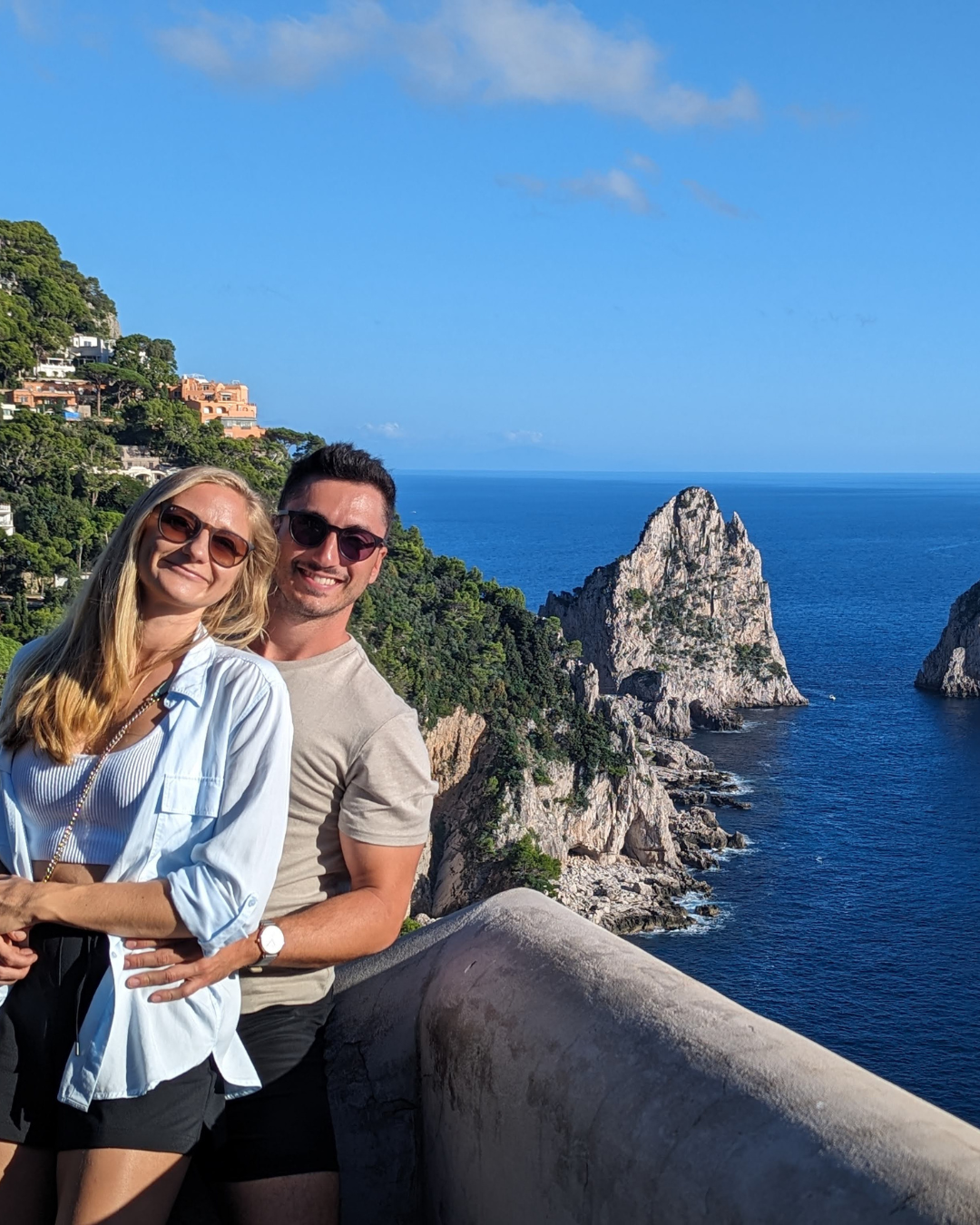 A couple standing close together on a scenic seaside overlook with cliffs and the ocean in the background, smiling and wearing sunglasses.