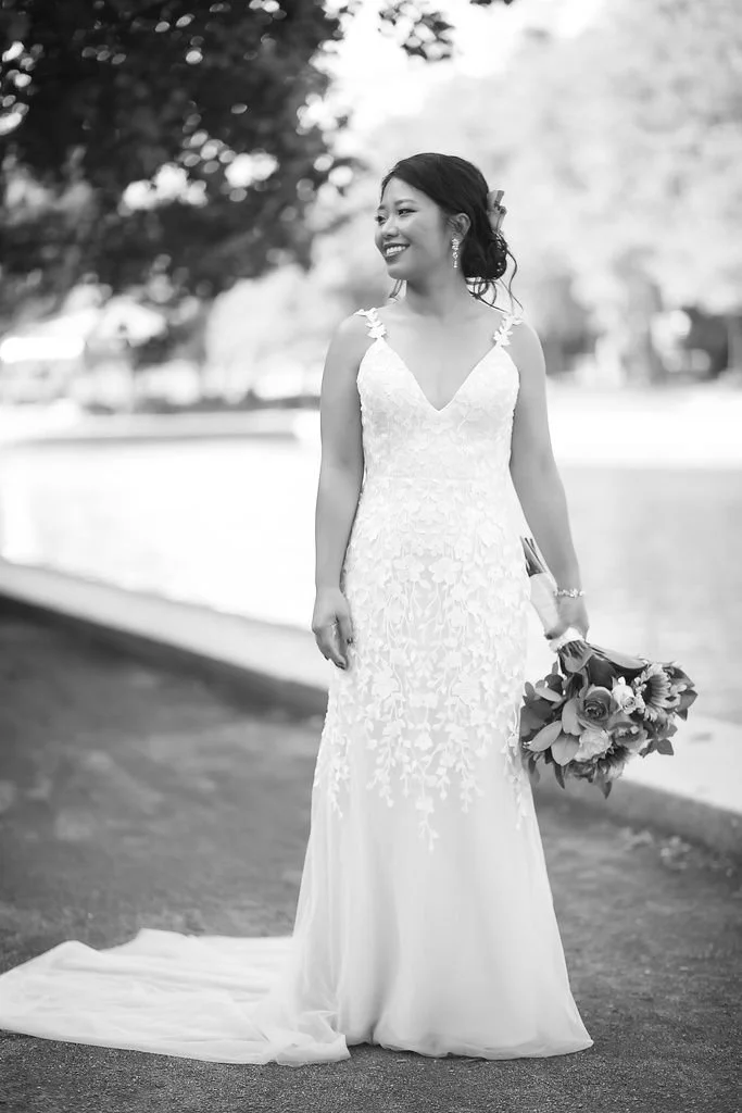 A woman in a wedding dress holding a bouquet, standing outdoors on a sidewalk, smiling, with trees in the background.