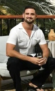 A man with a beard and short dark hair, smiling and sitting outdoors on a white cushioned chair, holding a glass of beverage. He is wearing a white short-sleeve shirt and black pants, with a yellow object on a small white table beside him. Background includes green foliage and a black railing.