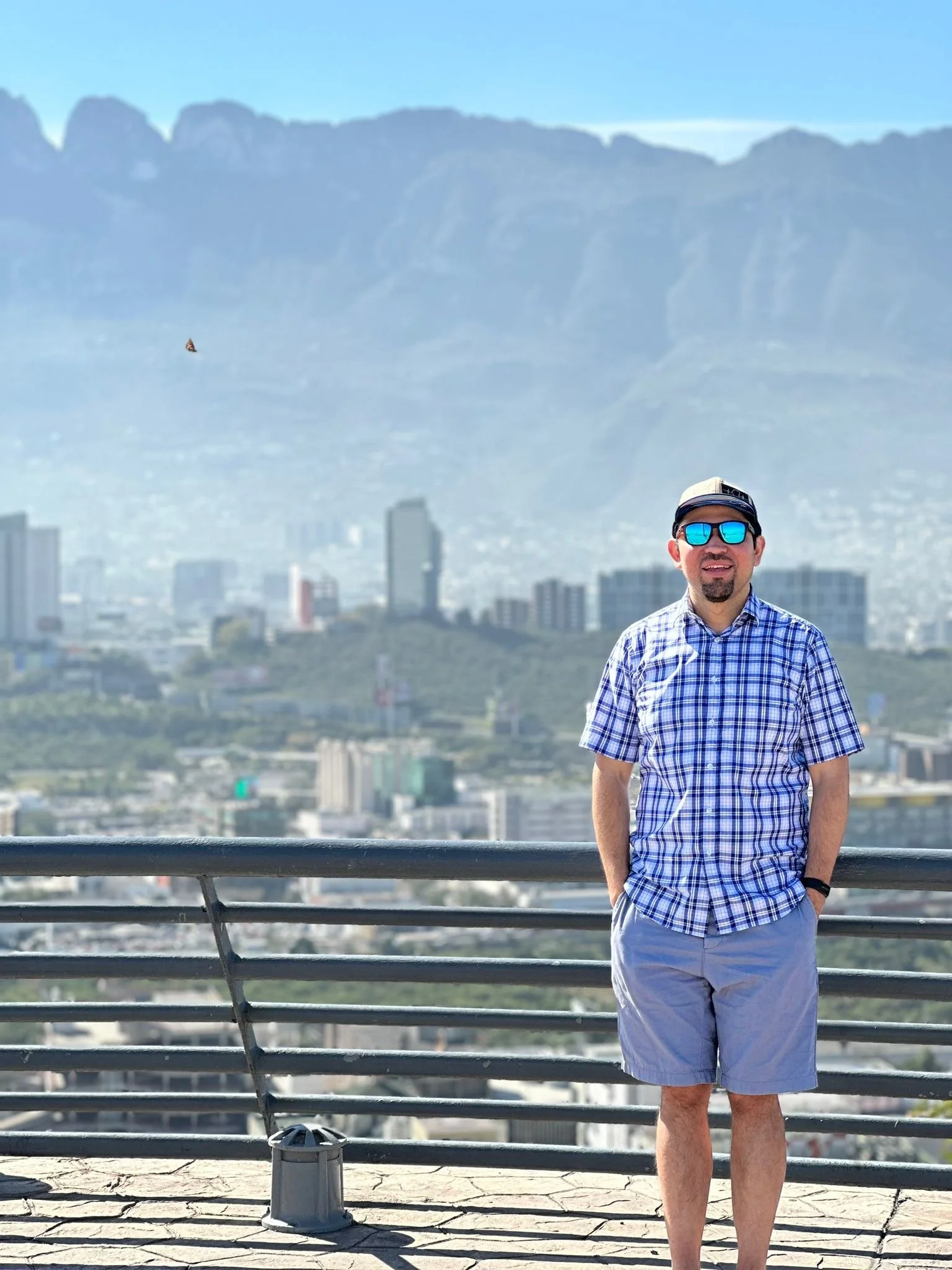 A man wearing a plaid shirt, shorts, sunglasses, and a cap standing on a balcony with a cityscape and mountain range in the background.