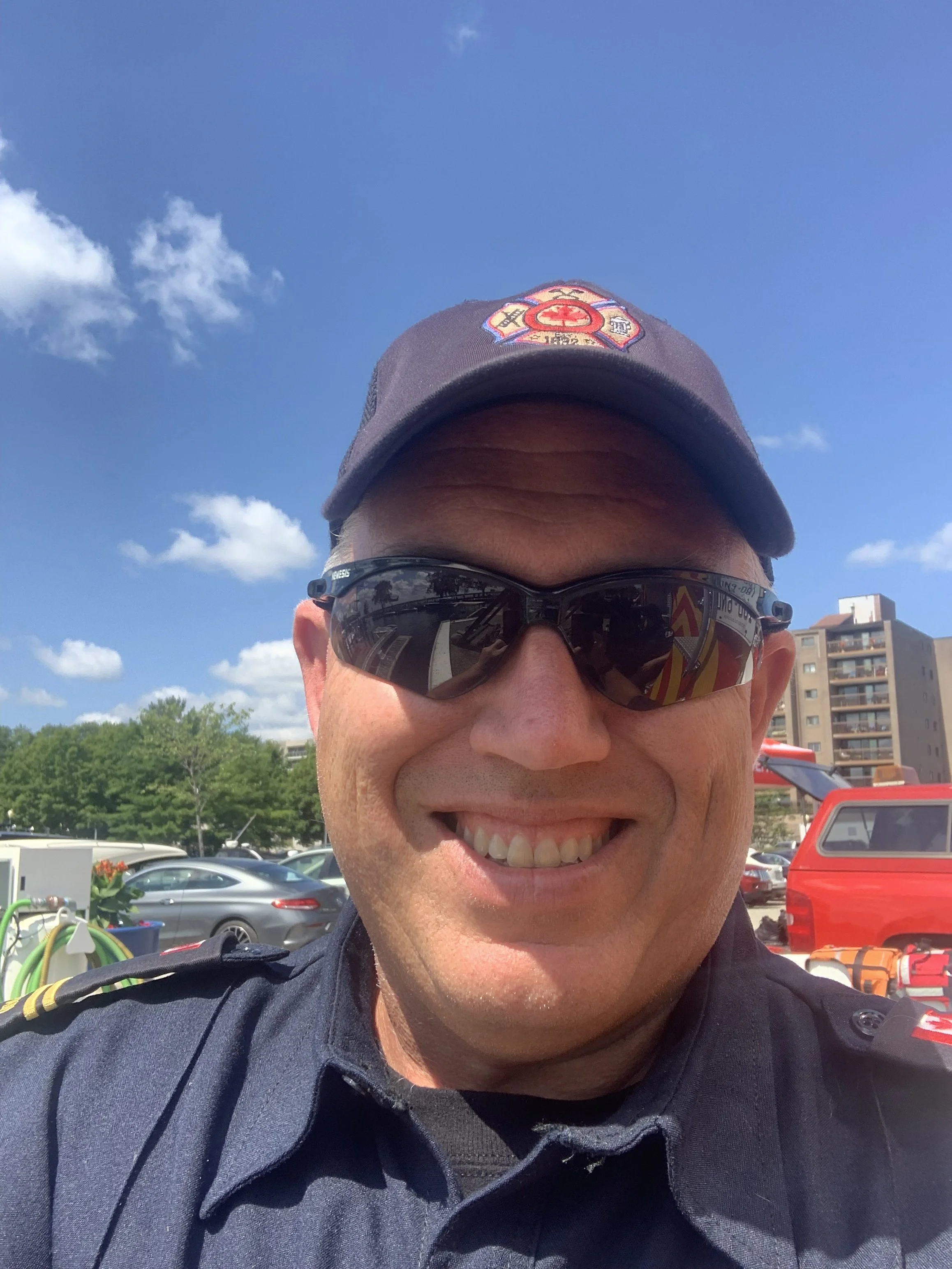 A smiling man in a firefighter uniform wearing sunglasses and a cap with a fire department emblem, outdoors with blue sky and buildings in the background.