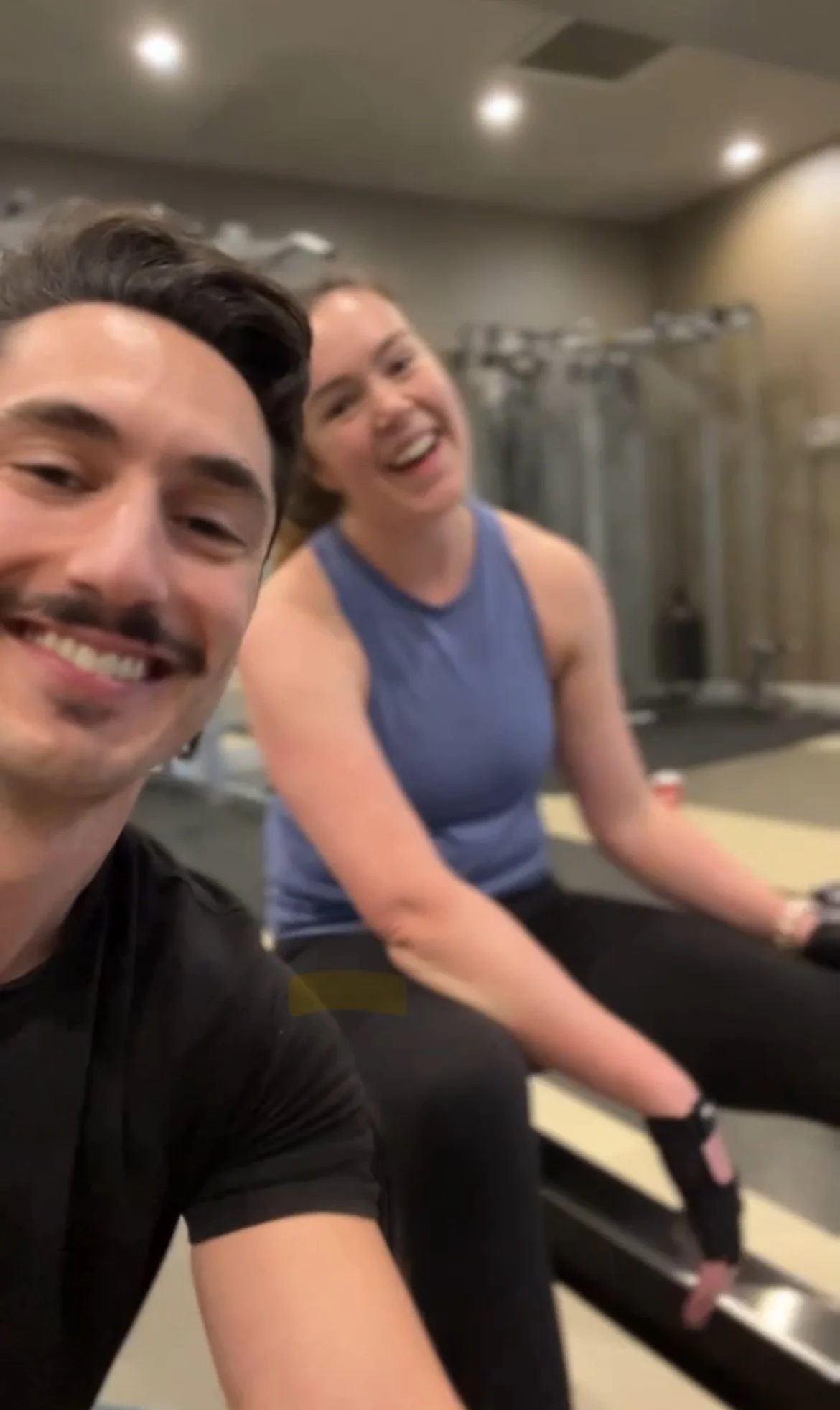 Two smiling people taking a selfie in a gym. The person on the left is a man with dark hair and a mustache, wearing a black shirt. The person on the right is a woman with brown hair, wearing a blue workout top, sitting on an exercise bench with workout equipment in the background.