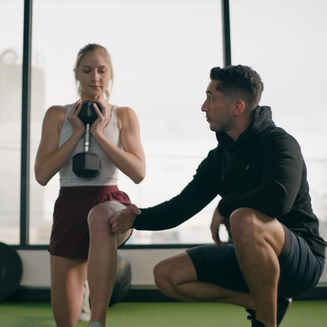 A woman holding a dumbbell while a male trainer assists her with her knee during a workout session in a gym.
