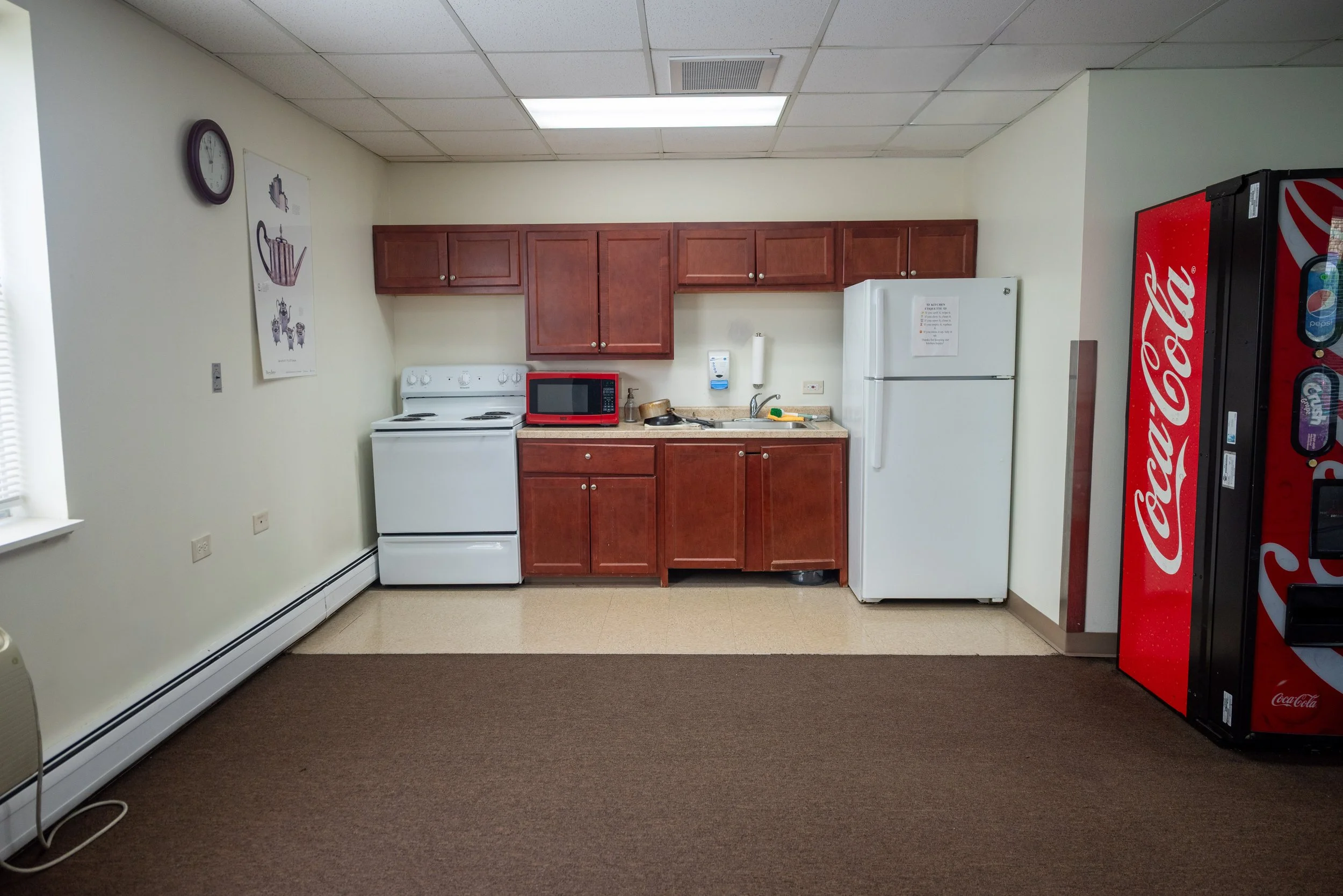 A small kitchenette with wooden cabinets, a white refrigerator, a red microwave, a sink, and a Coca-Cola vending machine against a plain white wall with a clock and a poster.