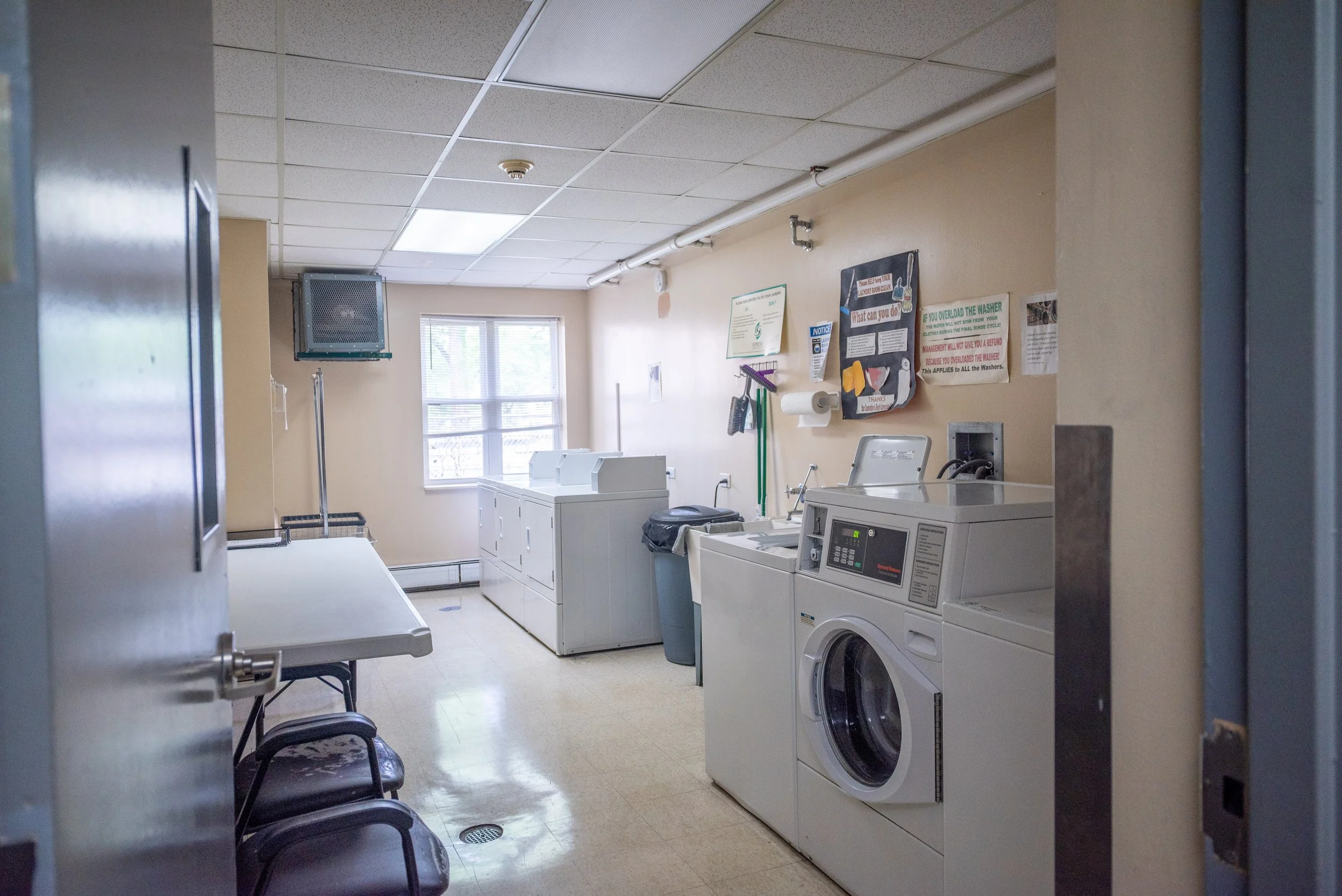 A laundry room with washing machines, a laundry basket, a trash can, chairs, and informational posters on the wall.