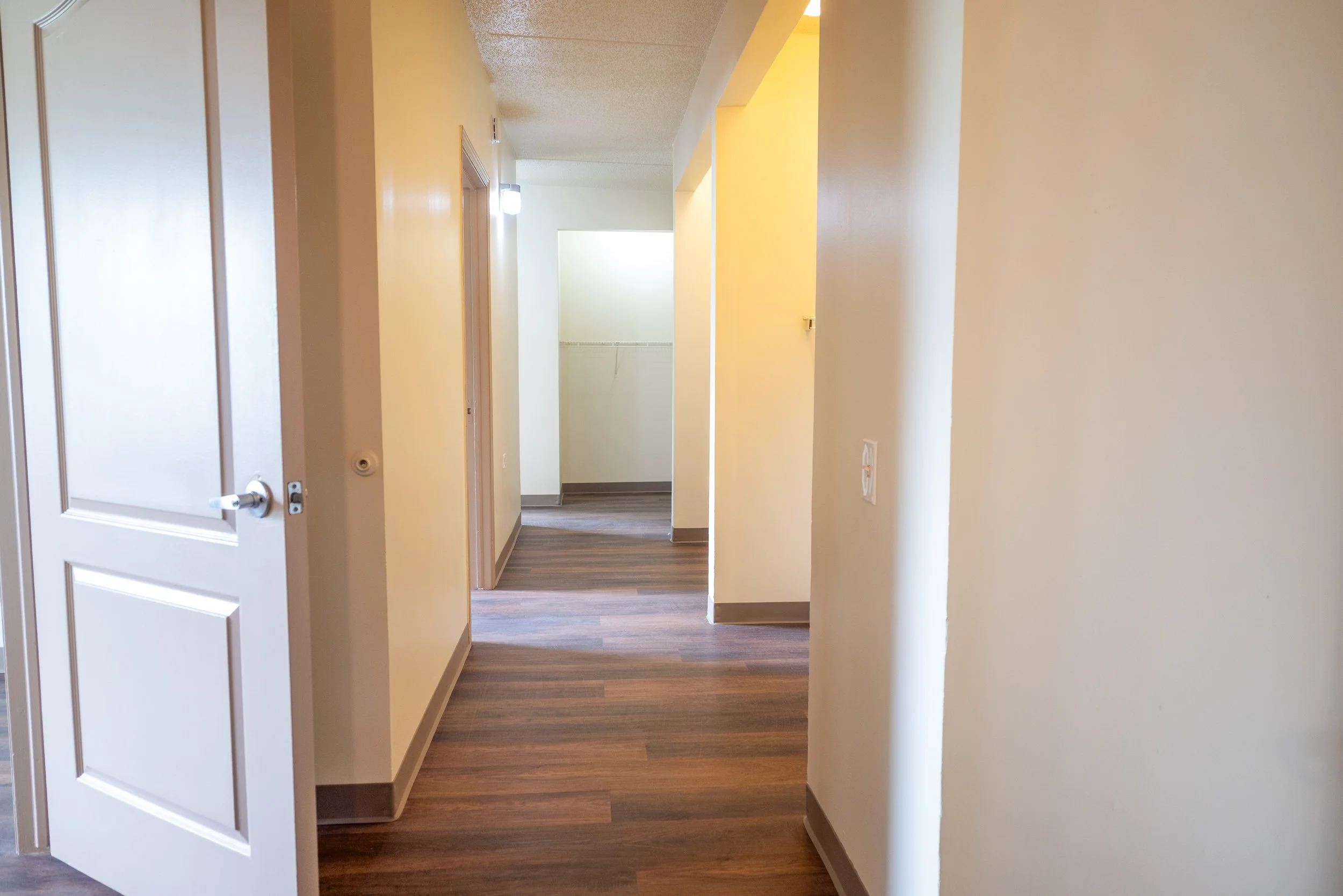 View of an empty hallway with cream-colored walls, wooden flooring, and an open closet at the end.
