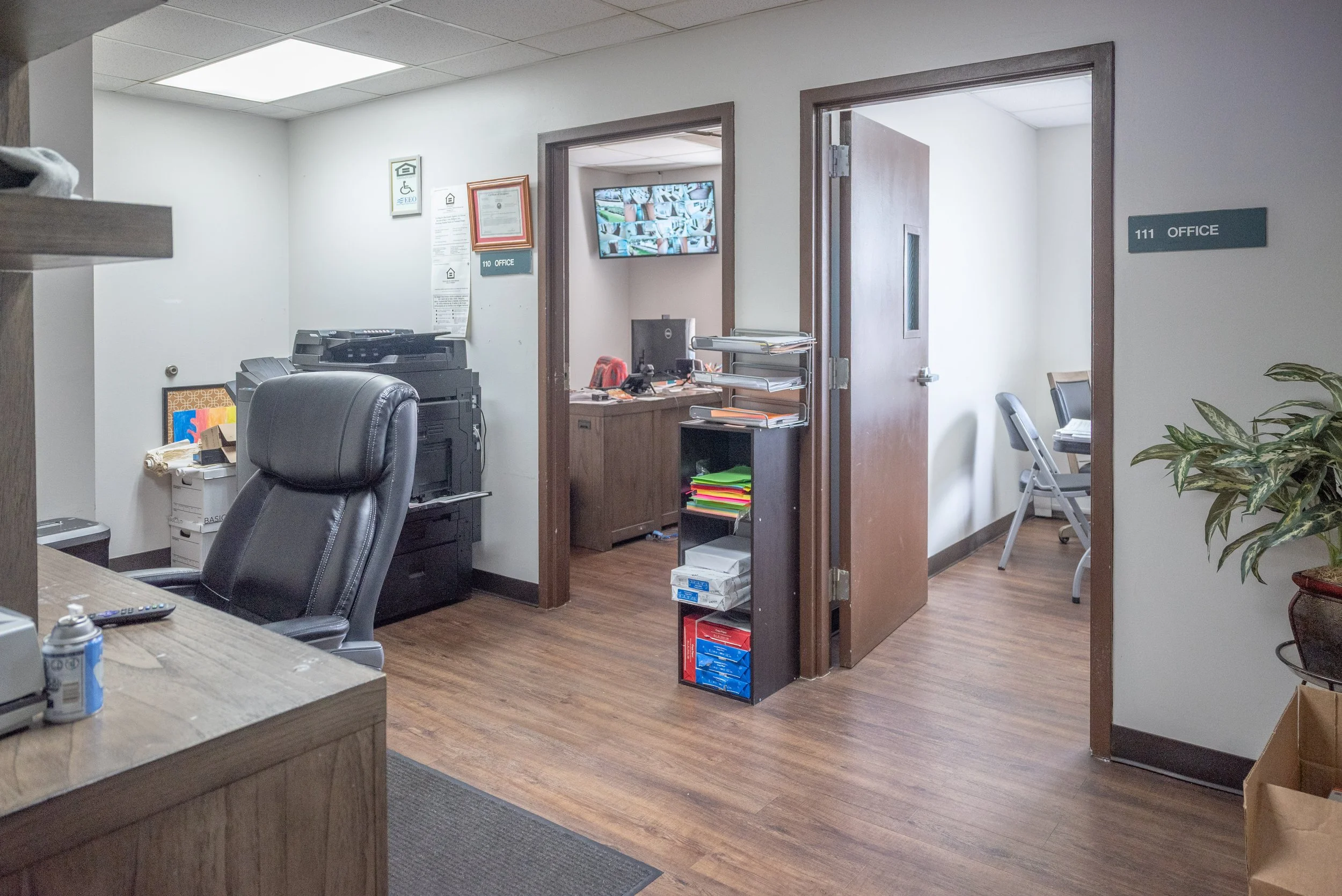 Office area with desks, chairs, a computer monitor displaying security camera feeds, a printer, shelves with office supplies, and doors labeled 110 and 111.