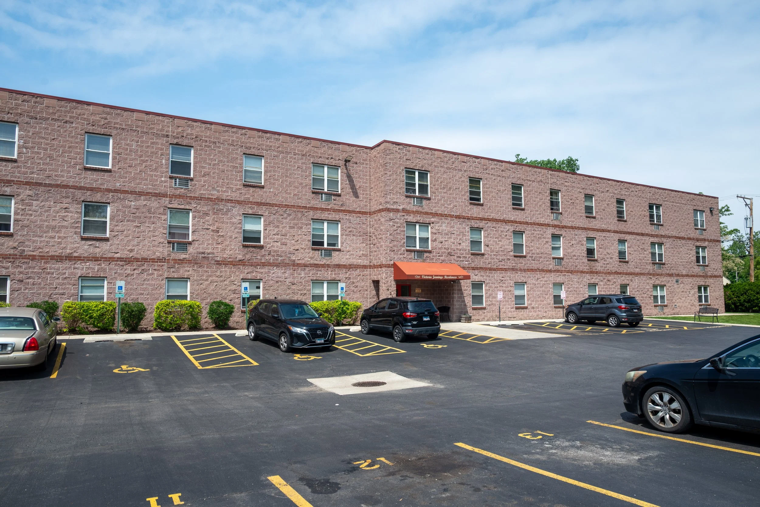 Parking lot with six cars parked in front of a three-story pink brick apartment building with six rows of windows and an orange awning at the entrance. The parking spaces are marked with yellow lines; some are designated for handicapped parking.