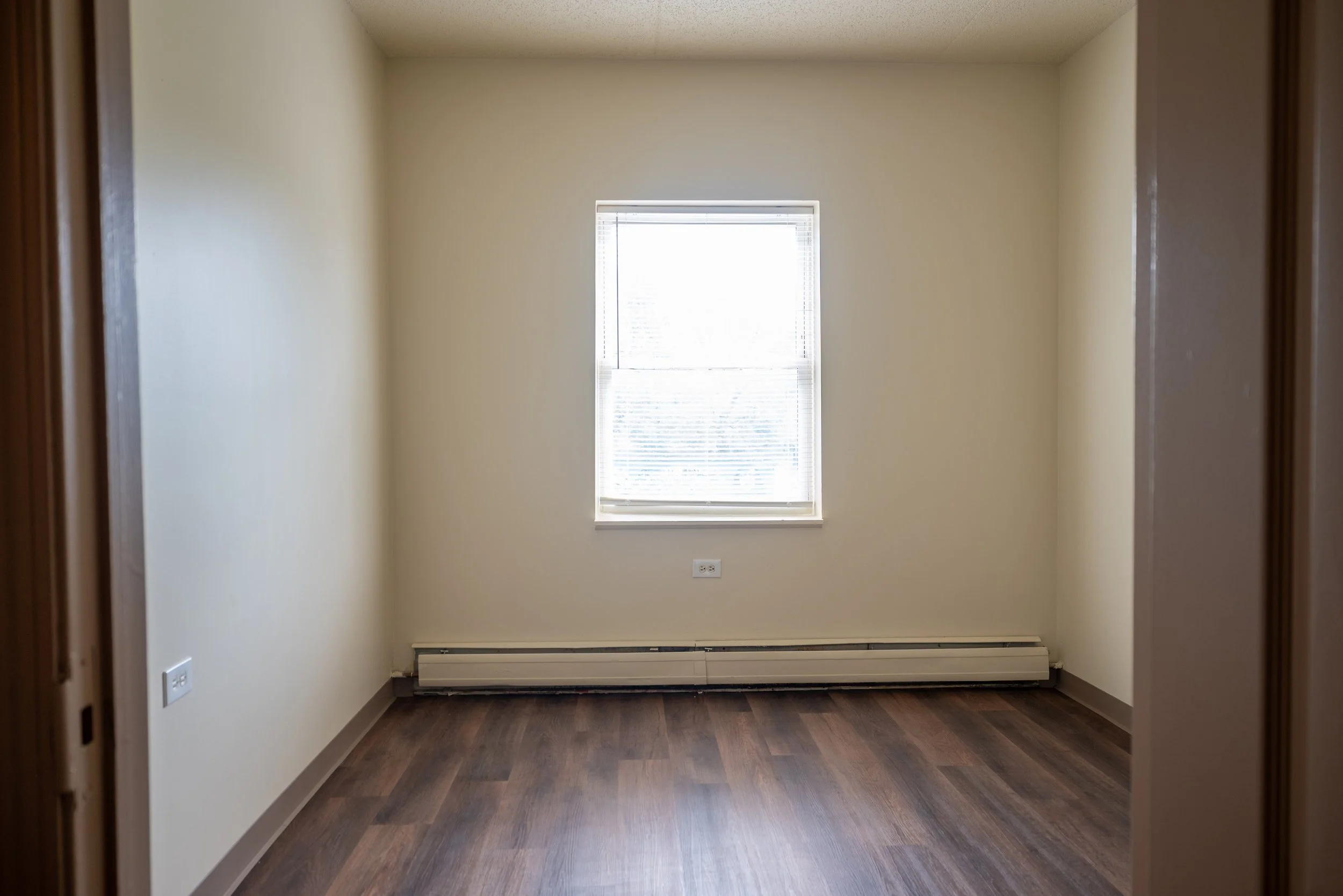 An empty room with beige walls, a window with blinds, and a dark wood floor.