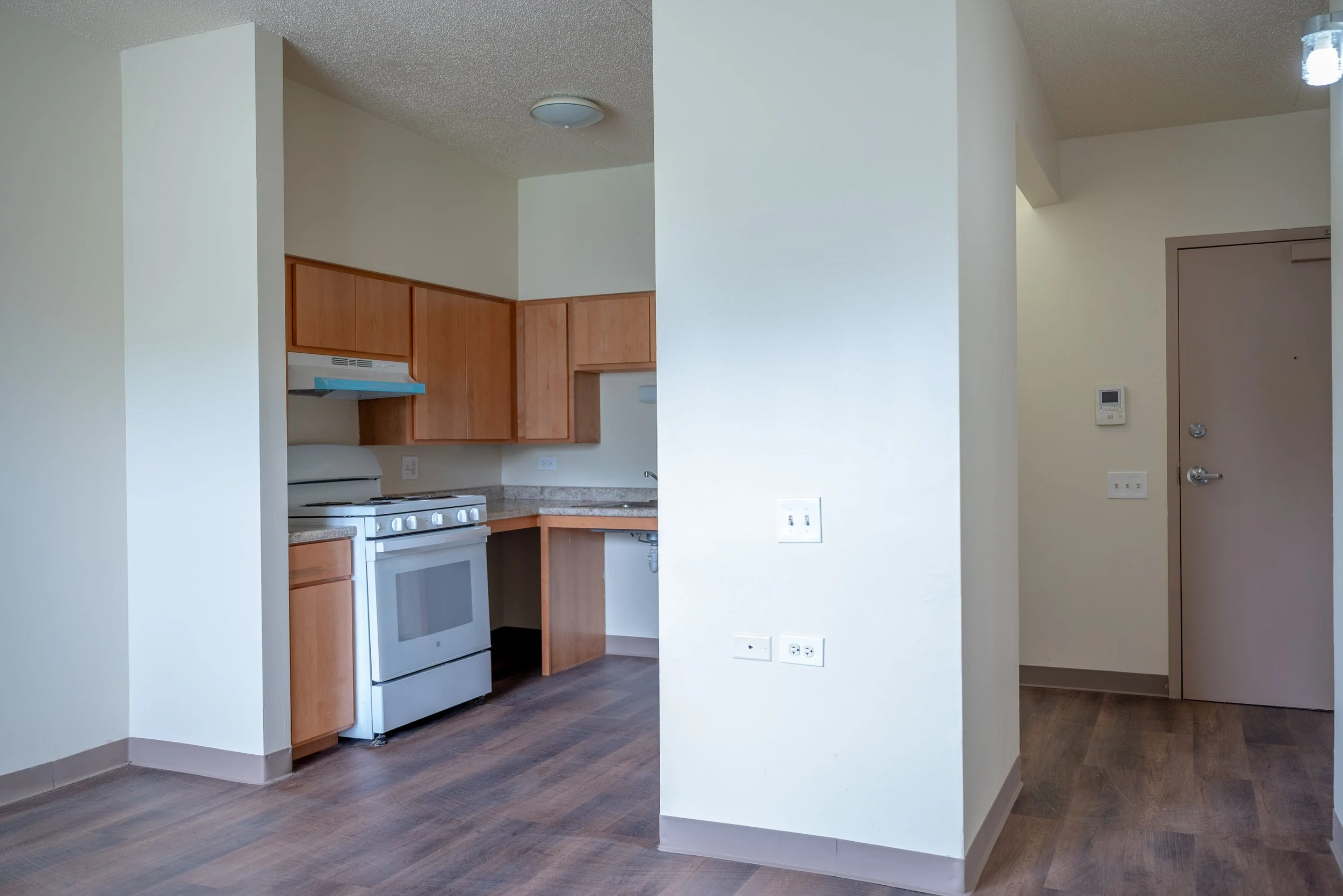 Part of a kitchen with wooden cabinets and a white stove, partially visible through a large opening in a white wall.