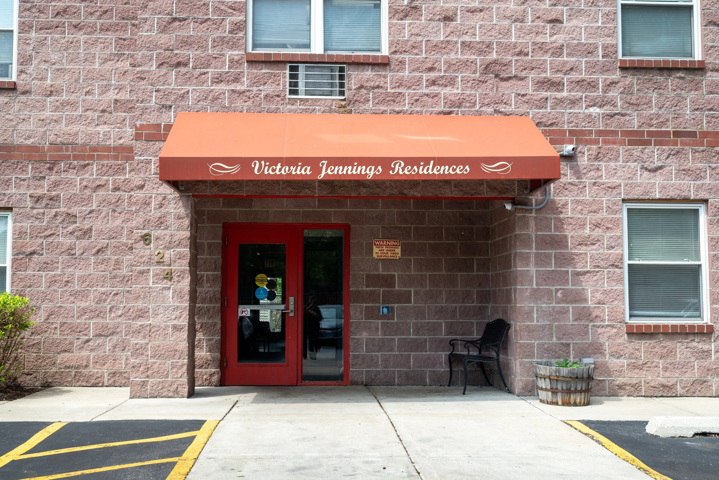 Entrance to Victoria Jennings Residences with a red door and a red canopy sign with white cursive writing. The building has a pinkish stone exterior, and a small bench sits to the right of the door. There is parking with yellow lines marked in front 