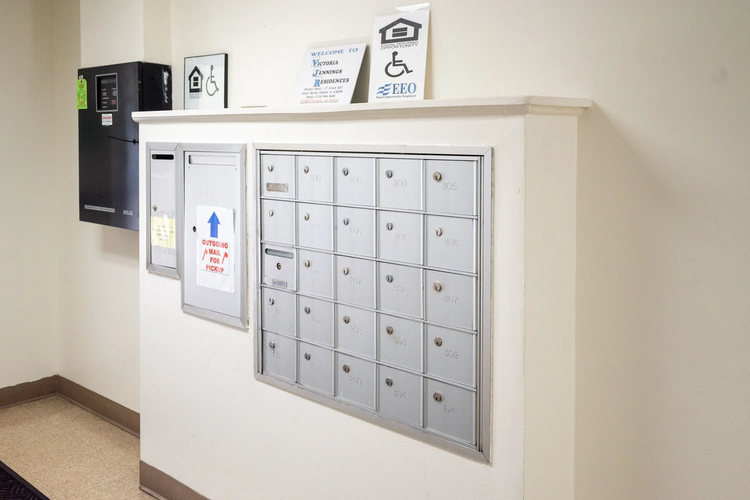 Wall-mounted mailboxes and a mailbox for outgoing mail in a hallway with beige walls and carpeted floor.