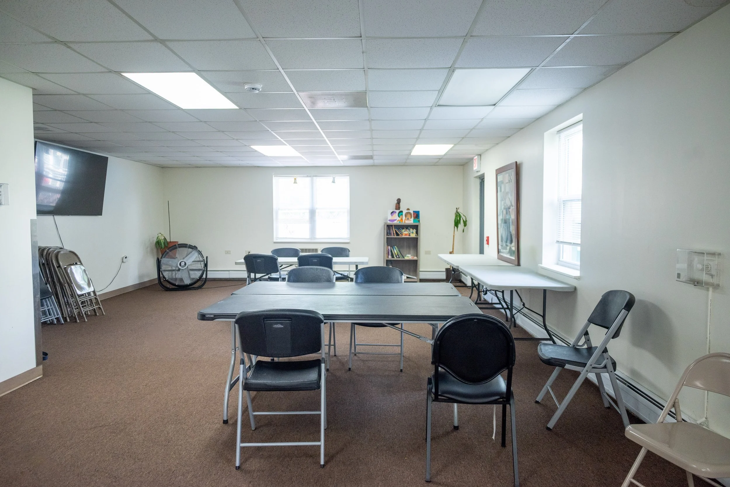 A conference room with tables and black chairs, large windows, a bookcase with books and decorations, a large flat screen TV mounted on the wall, and folding chairs stacked in a corner.