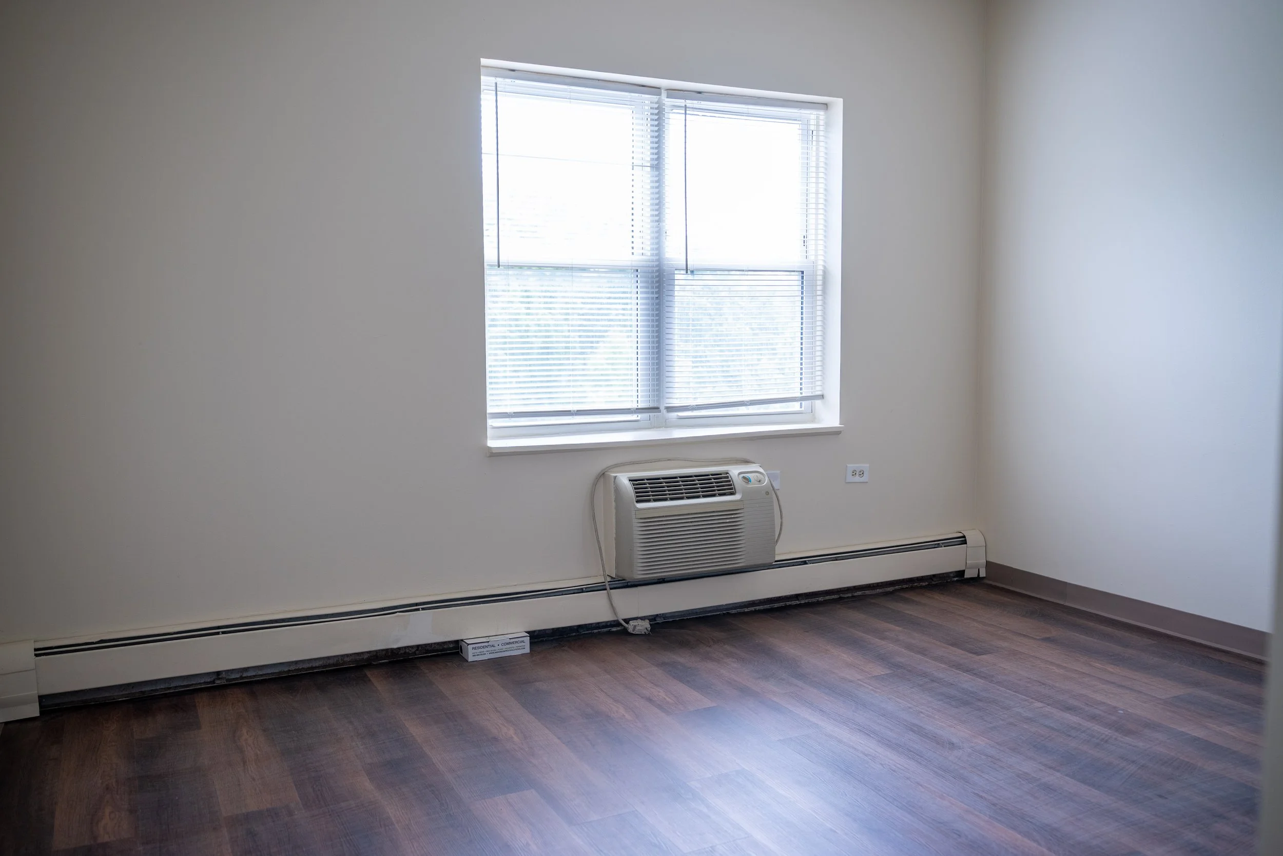 Empty room with a window, an air conditioner below the window, and a heating baseboard along the wall, with a power outlet and a small white box on the floor.