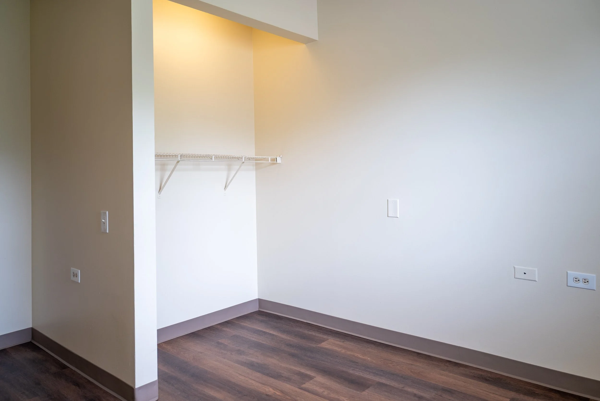 Empty room with white walls, dark wooden floors, and multiple electrical outlets. A small section of wall has a white wire shelf and is illuminated by a ceiling light.