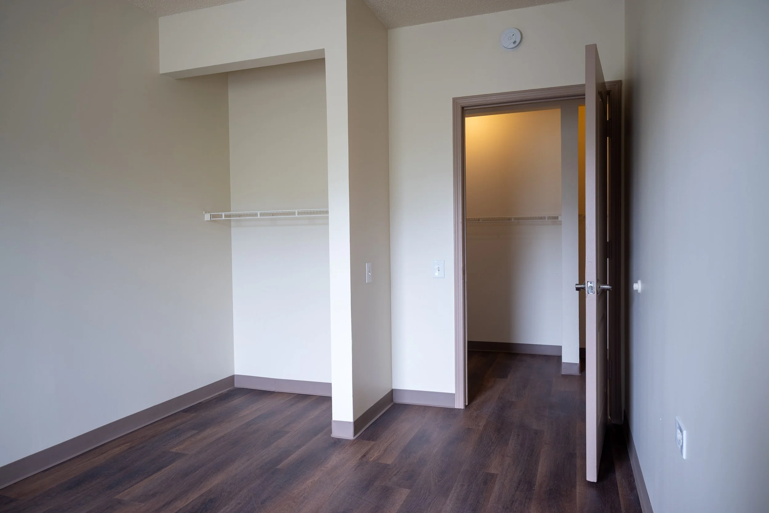 Empty room with beige walls, dark wood flooring, an open closet with a wire shelf, and a doorway leading to another room.