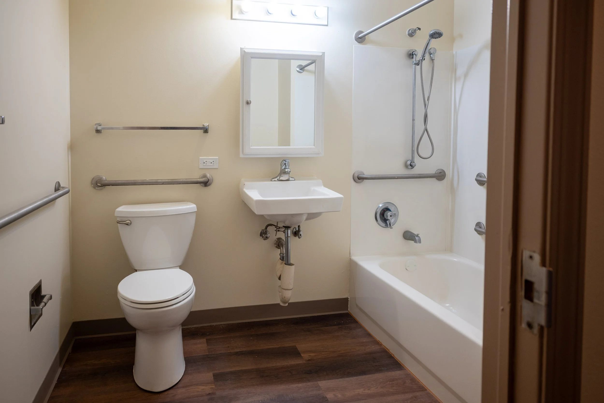 A compact bathroom with a white toilet, a small white sink, and a shower with grab bars. There is a mirror above the sink and a window above the shower. The bathroom has beige walls and dark wood flooring.