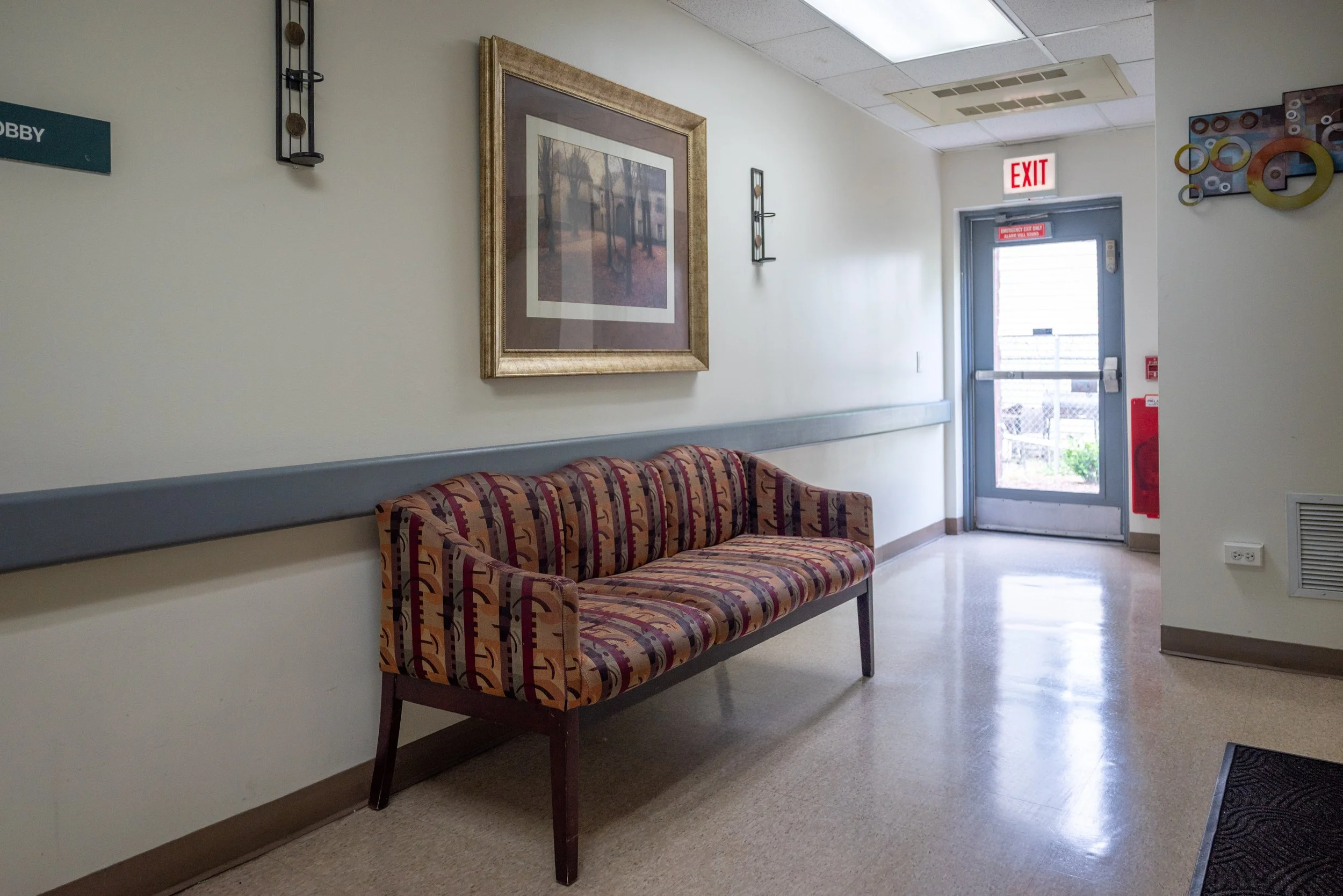 A waiting area in a building with a striped upholstered bench against a wall, a large framed picture of trees and a building, and a door with an illuminated exit sign above it.