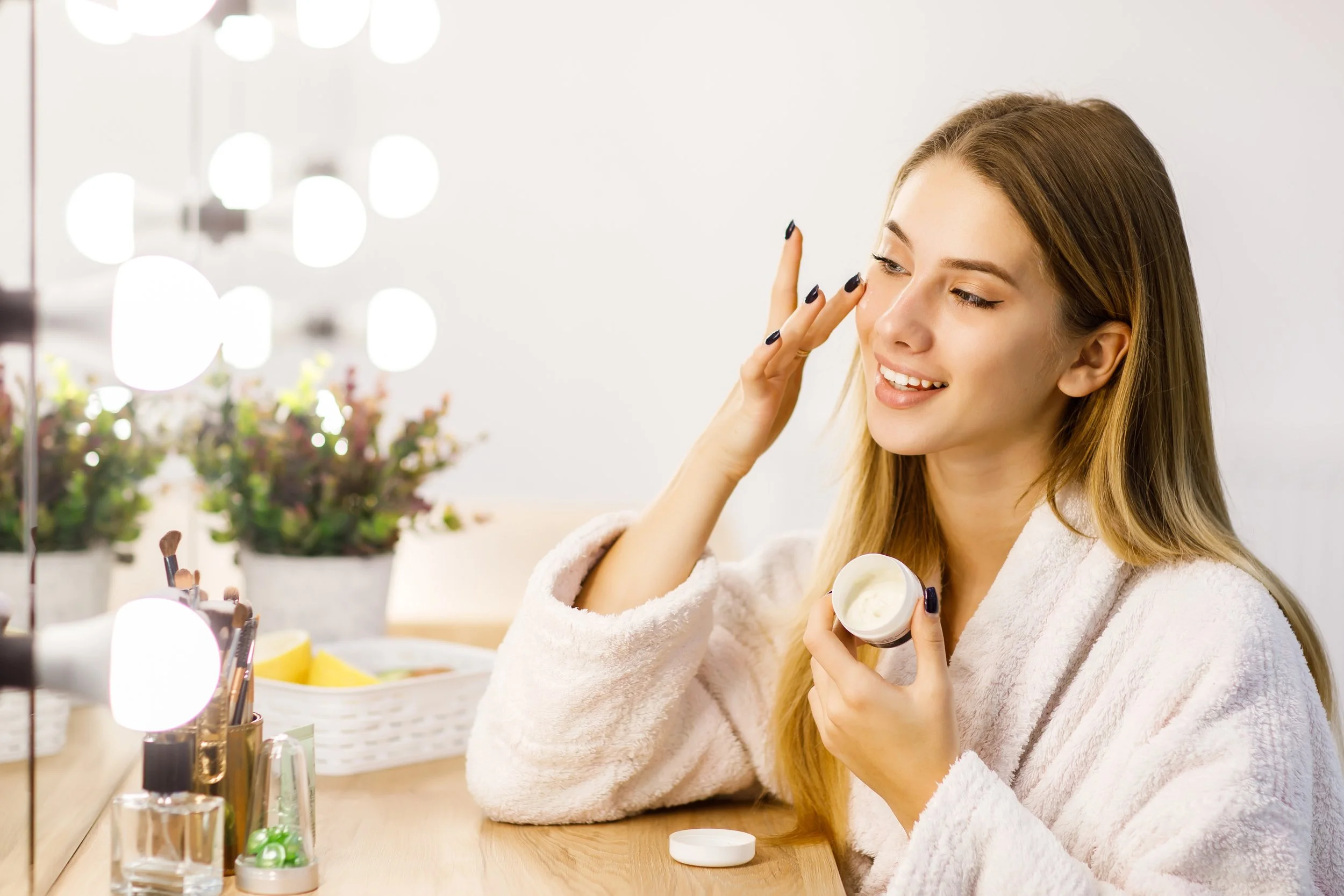 A woman with long blonde hair smiling and applying cream to her face in front of a mirror with bright vanity lights, surrounded by makeup brushes and beauty products on a vanity table.