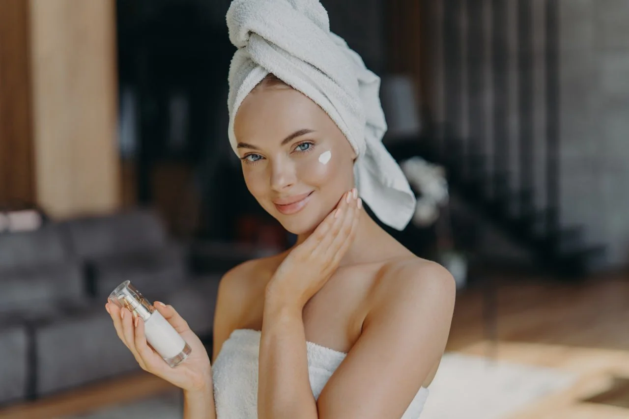 Young woman with a towel wrapped around her head applying skincare cream to her face, holding a small jar of moisturizer in her hand, in a cozy indoor setting.