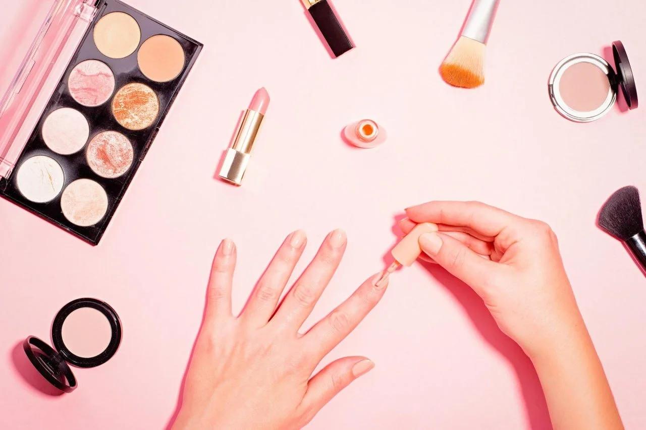 Person applying pink nail polish on their fingernails with various makeup items scattered on a pink surface, including an eyeshadow palette, lipstick, blush, brushes, and a mirror.