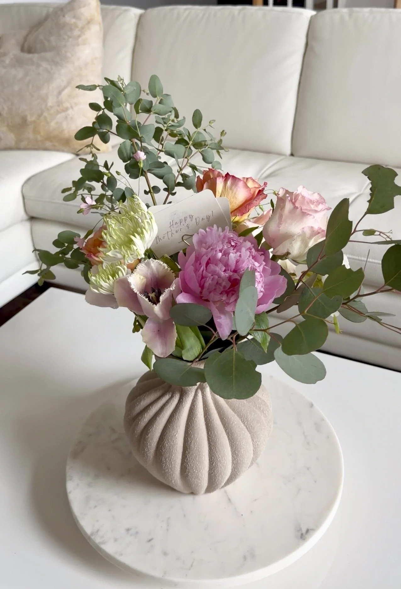 A beige, textured, round vase with ridges, containing a bouquet of pink and white flowers and green foliage, placed on a white marble stand with a marble surface beneath.
